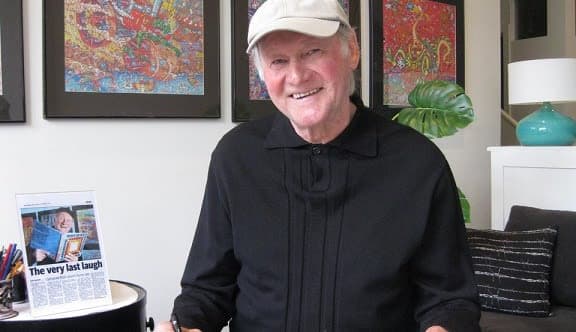 An older man in a living room with art on the walls smiles at the camera