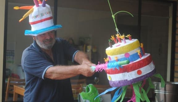  A smiling man, wearing a hat in the shape of a birthday cake, takes a swing at a birthday cake pinata
