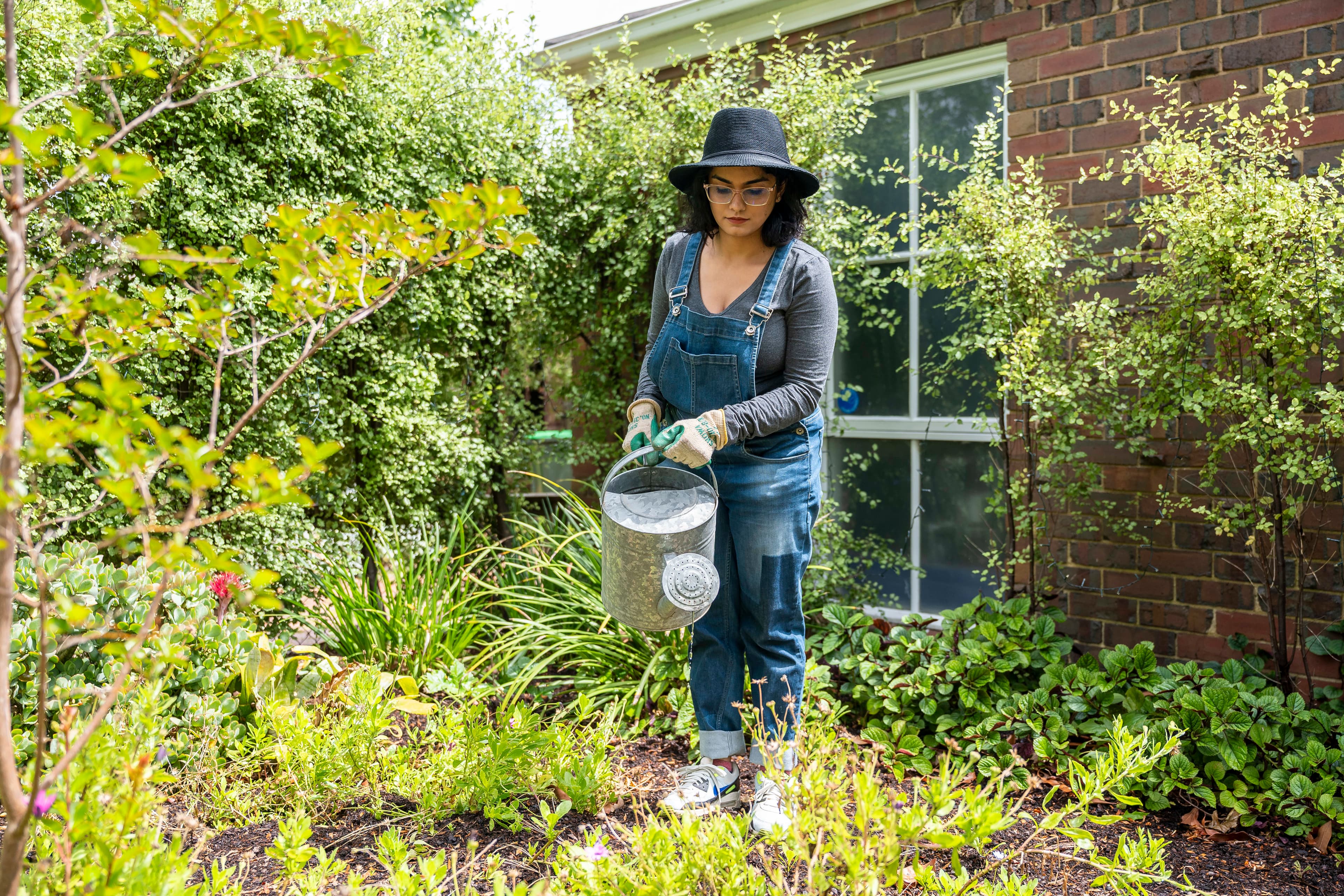 A woman stands in a garden outside a house holding a watering can and wearing denim overalls, a black wide-brimmed hat and gardening gloves.