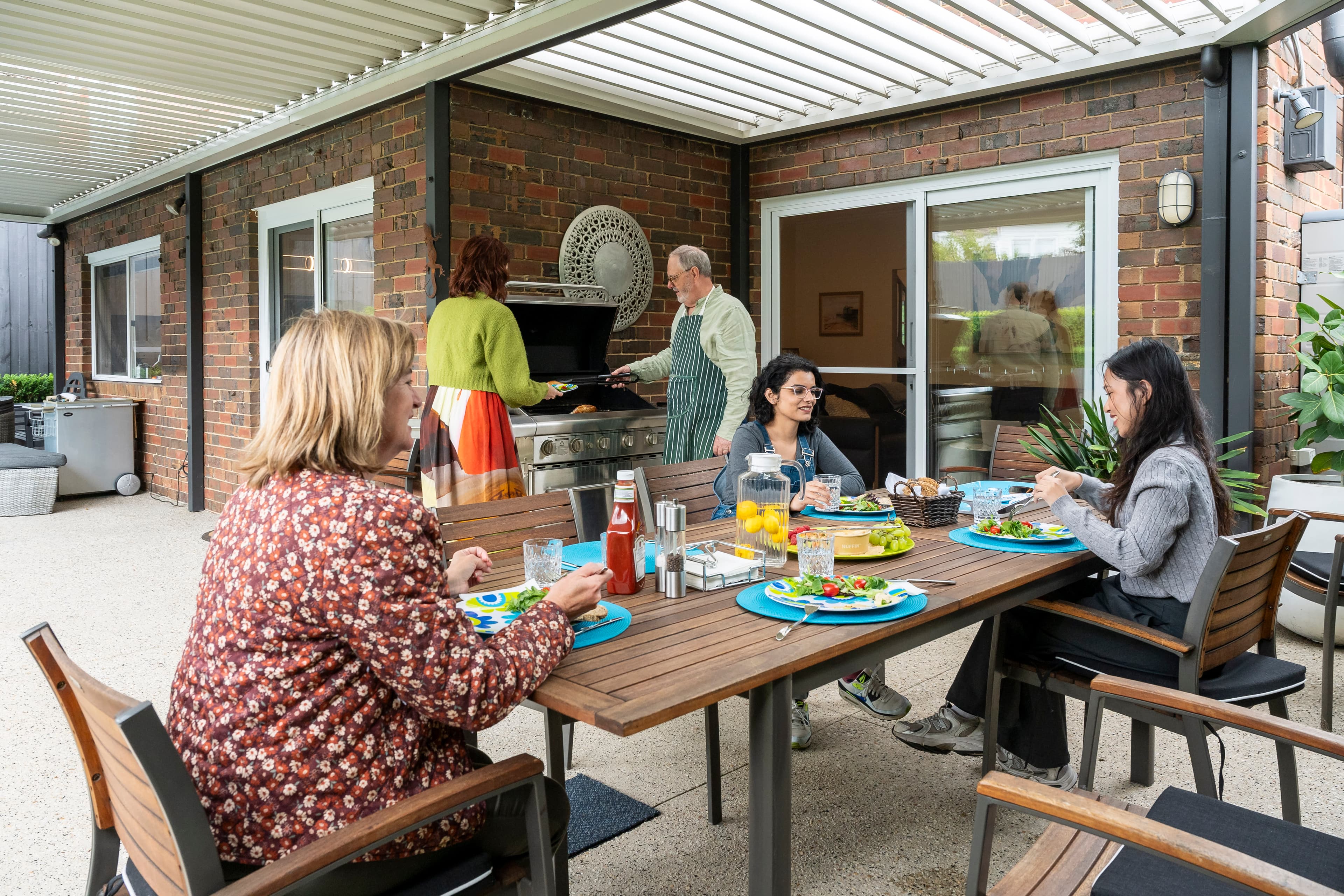 Five adults gather for a barbecue with three siting at an outside table and two others standing at a barbecue.