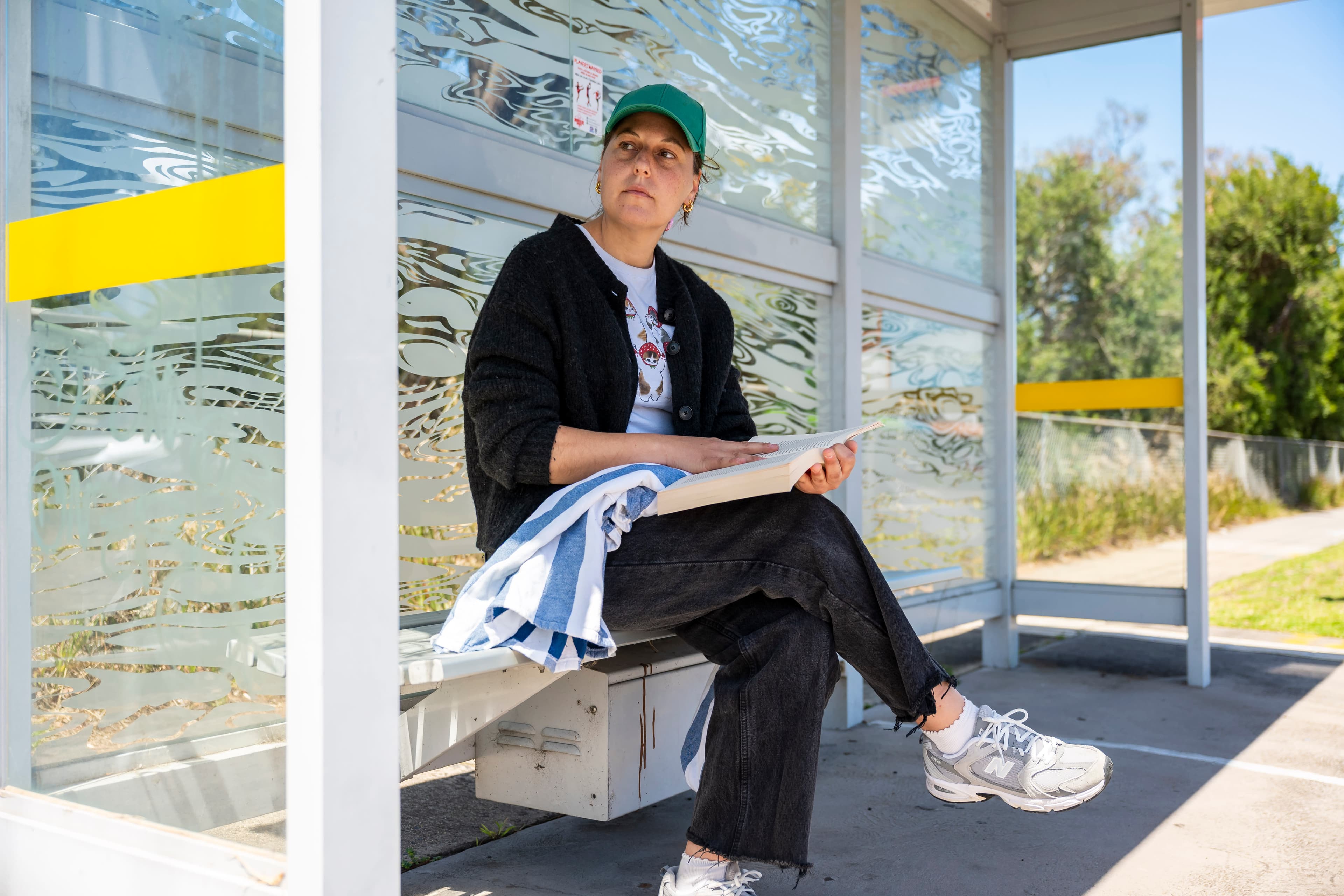 A woman in a baseball cap sits at a bus stop.