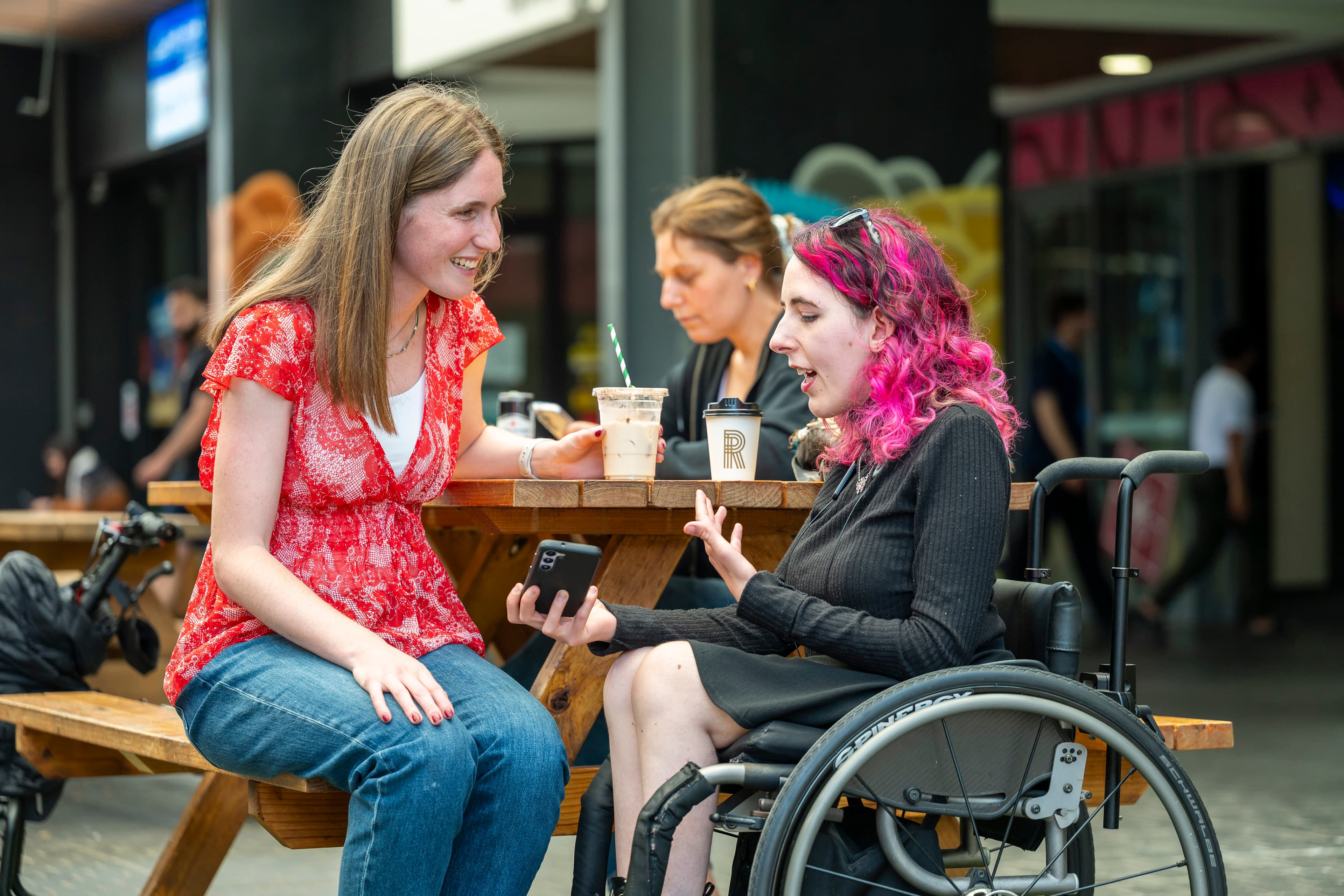Two young women at a shopping centre have coffee together and look at a phone