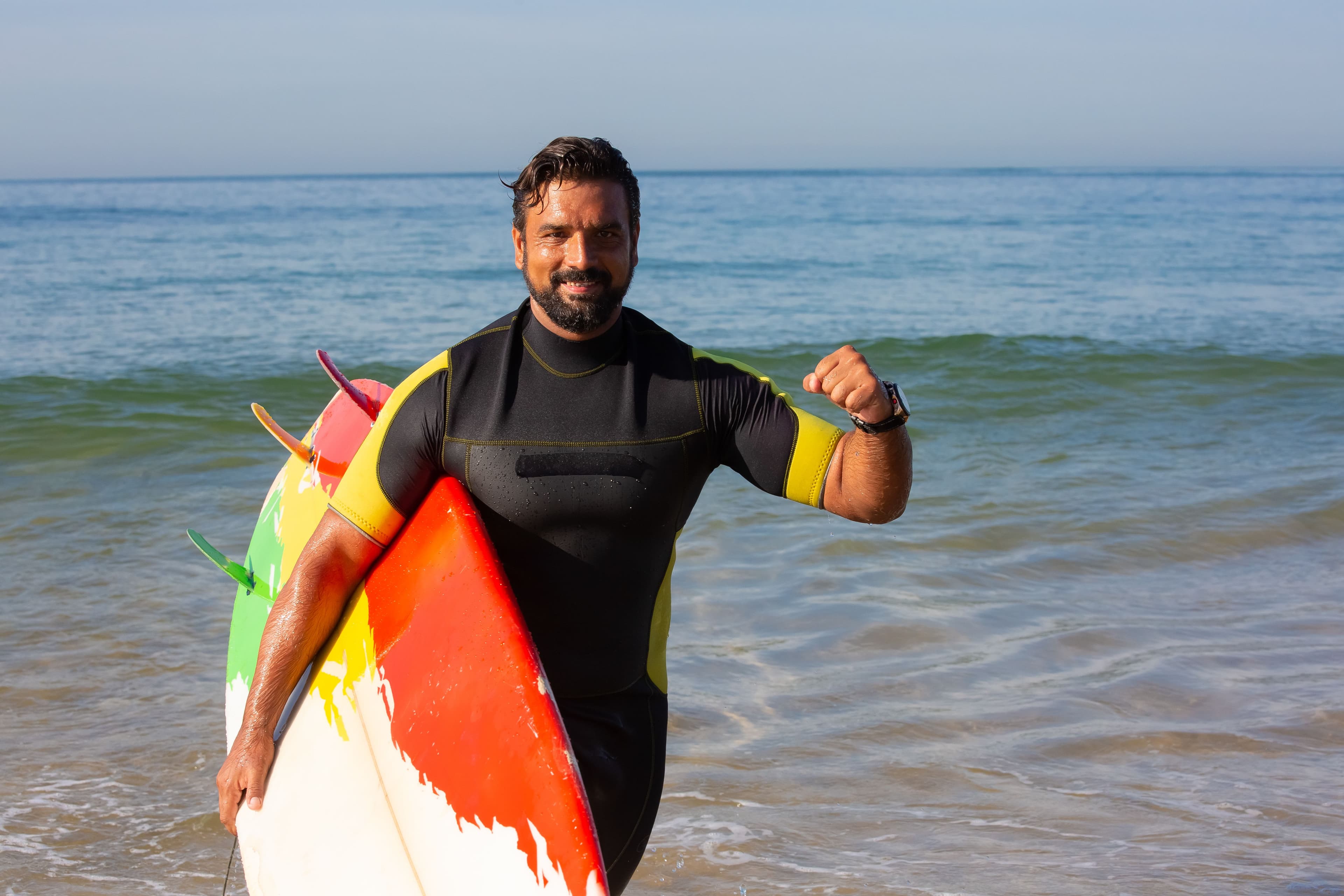 A surfer in a wetsuit carrying his board under one arm emerges triumphantly from the waves.