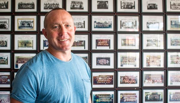 A man in a blue shirt stands in front of a wall of photos