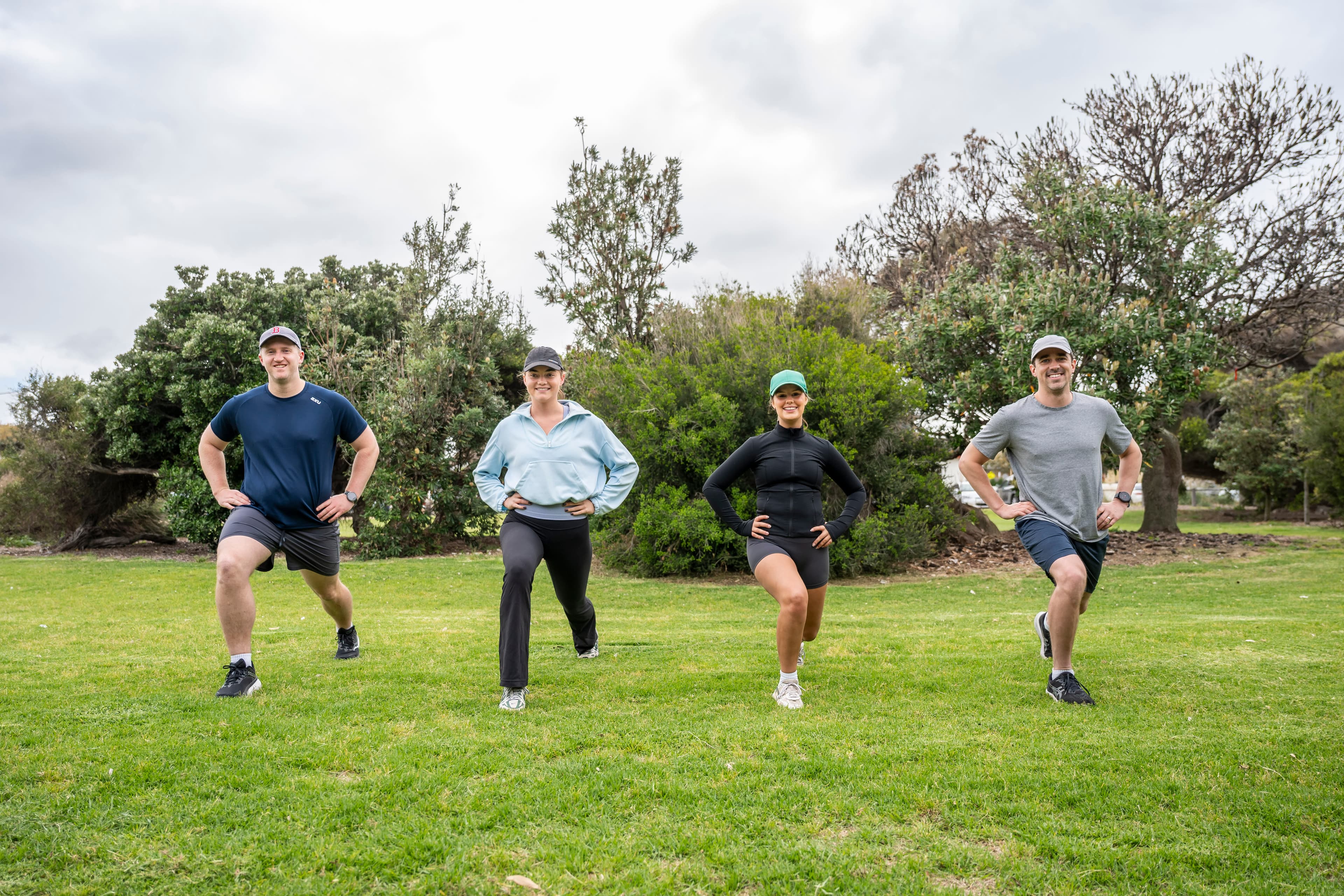 Four people who are exercising step forward in a stretch while standing on grass with trees behind them.