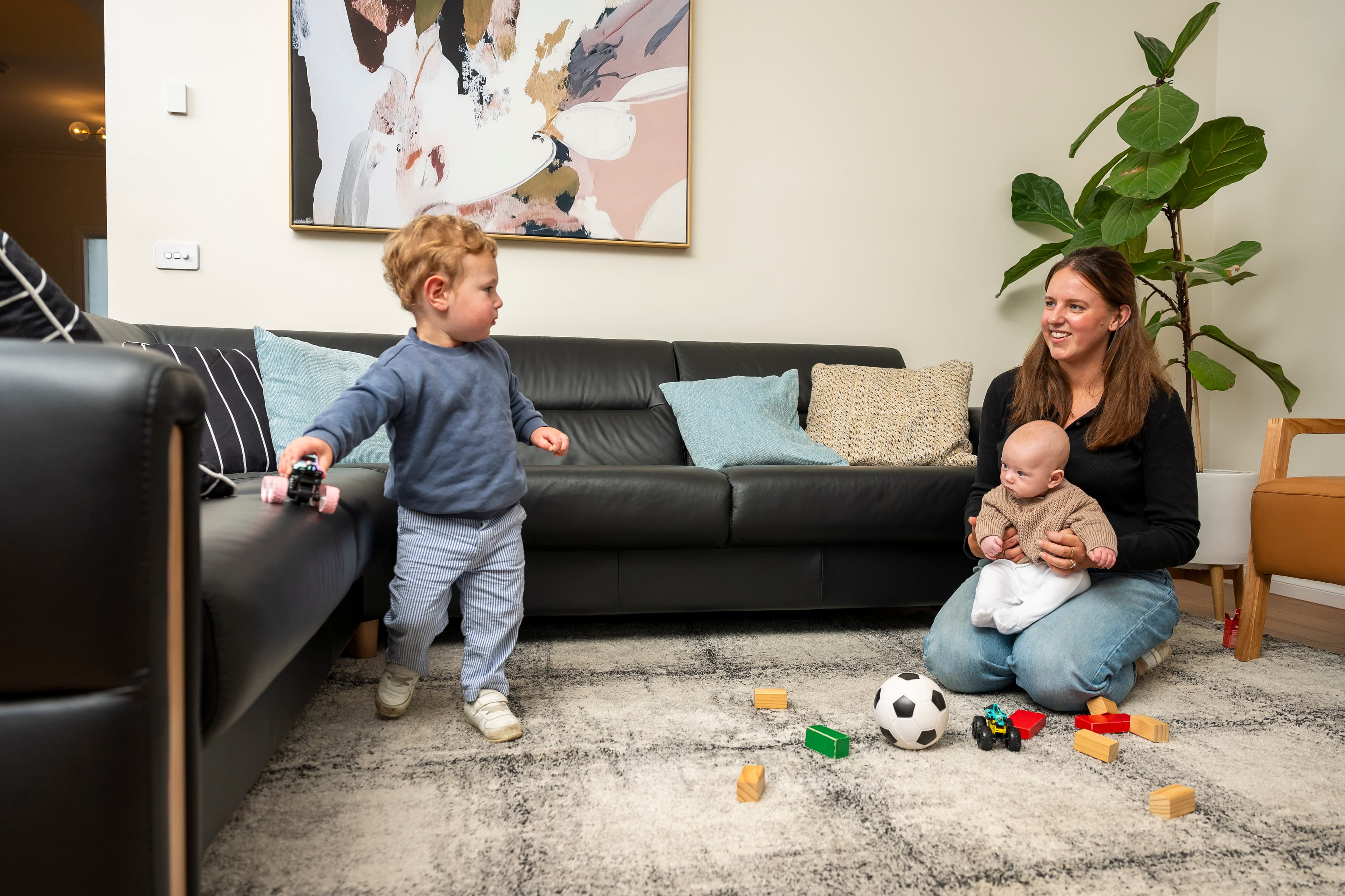 A woman sits on a rug holding a baby and looks at a toddler holding a toy and resting against a couch.