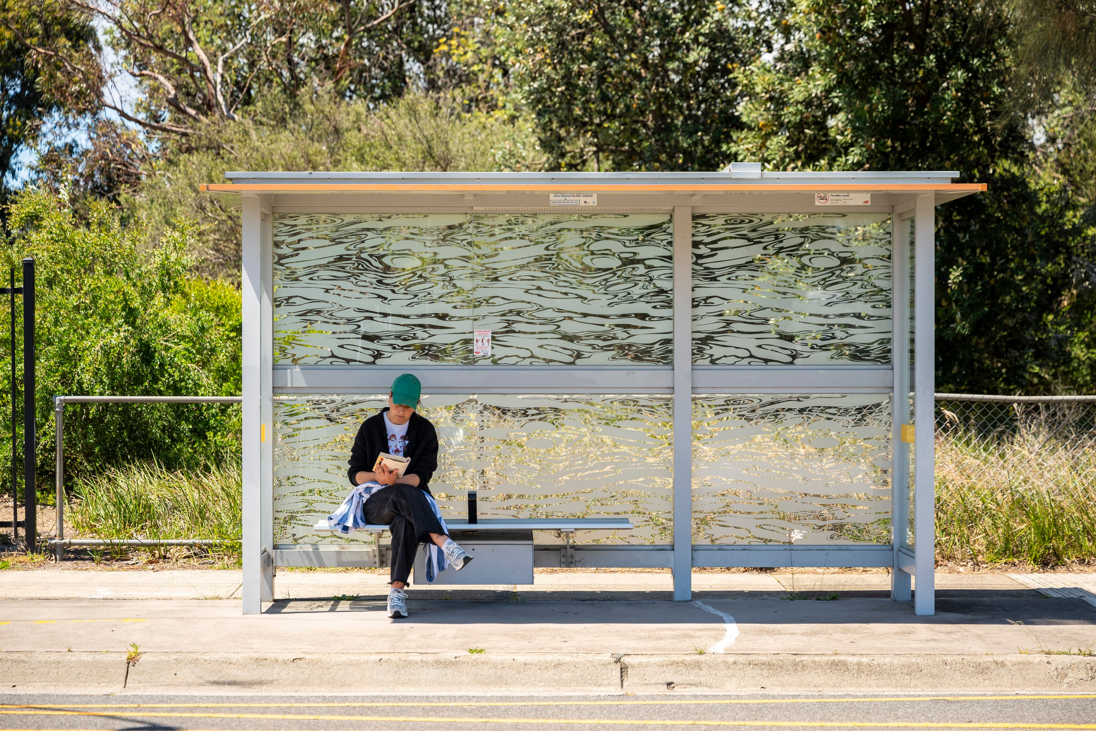 A person wearing a green cap, black pants, a white top, black jacket and sneakers sits at a bus stop reading a book.